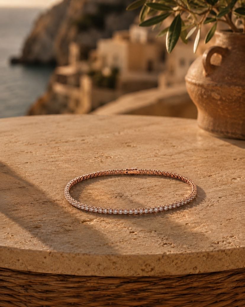 Bracelet tennis Valentina en plaqué or rose présenté en cercle au centre d'une dalle de travertin doré, alignement parfait des zircons taille brillant sertis griffes captant la lumière dorée du couchant, jarre en céramique terracotta avec olivier en haut à droite, village amalfitain perché sur les falaises et mer méditerranéenne en arrière-plan flou, panier en osier sous la dalle