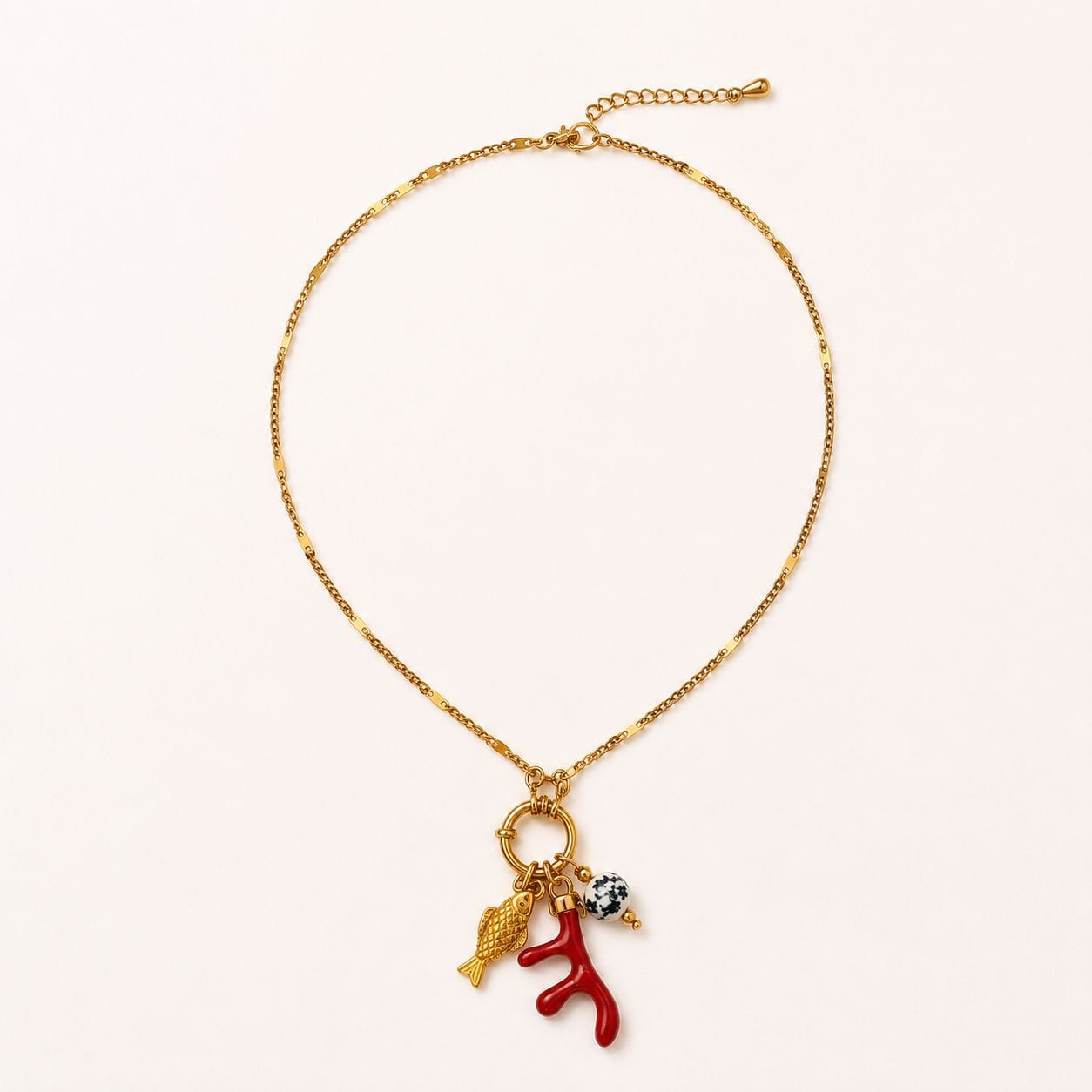 Collier Elina à plat sur fond crème pâle, chaîne fine à maillons alternés barre rectangulaire et maillon rond, cluster pendentif au bas — petit poisson d'or, brin de corail rouge méditerranéen, perle de jaspe dalmatien noir-et-blanc