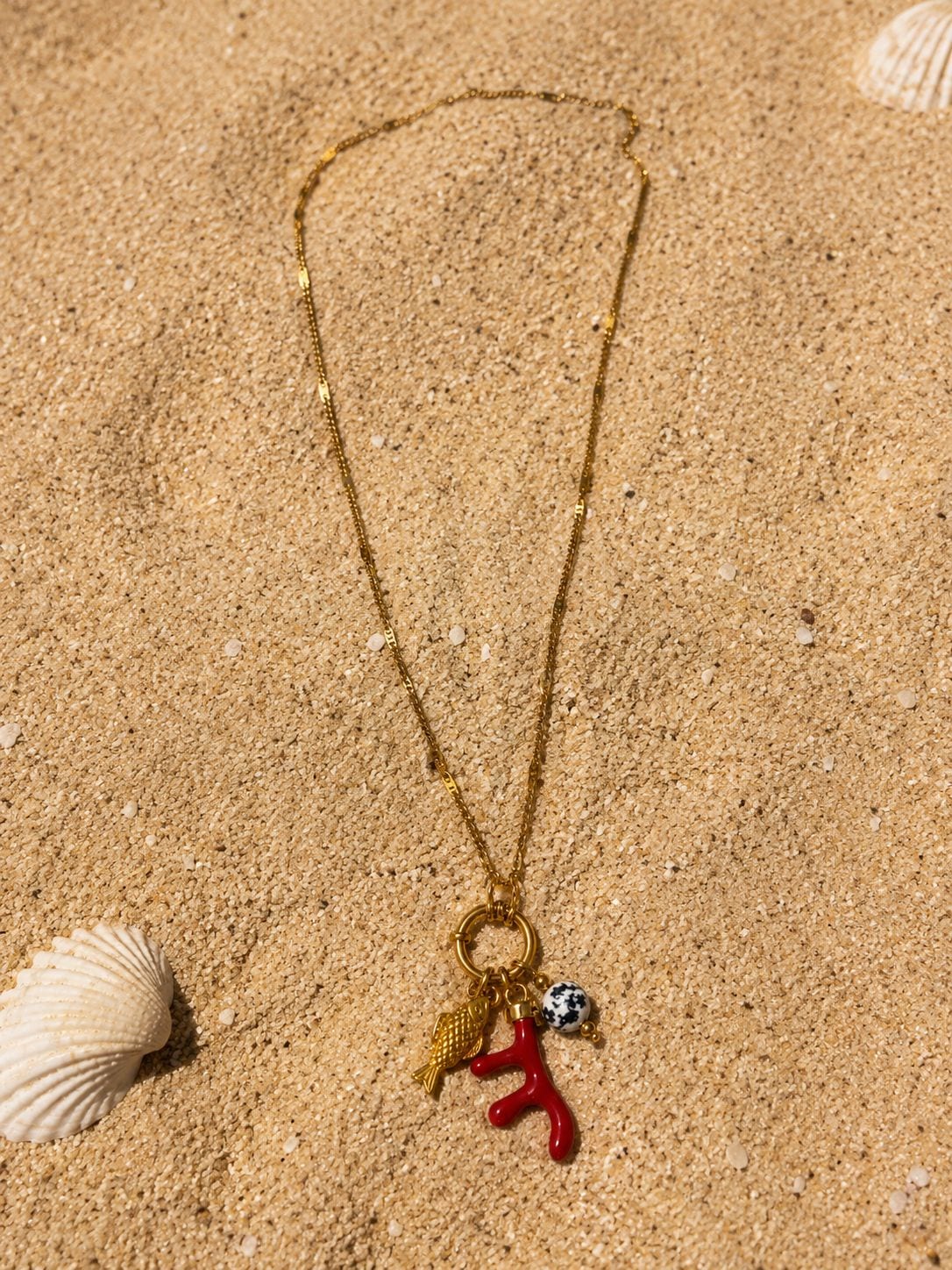 Collier Elina posé sur le sable doré, chaîne fine étirée en demi-cercle, cluster pendentif en bas et coquillage Saint-Jacques nacré à gauche, lumière du soleil zénithal