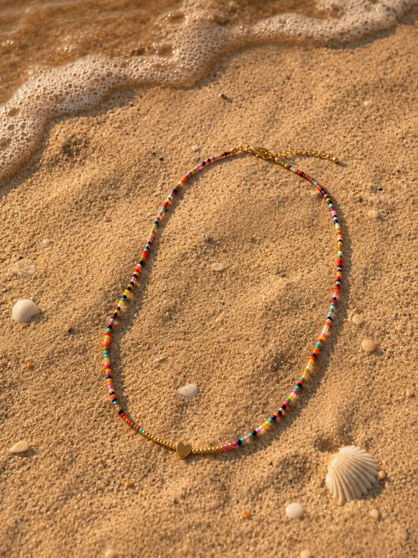Collier Elena posé sur le sable doré, étiré en demi-cercle, coquillages Saint-Jacques crème éparpillés autour, vague d'écume au loin, ombre de fin d'après-midi