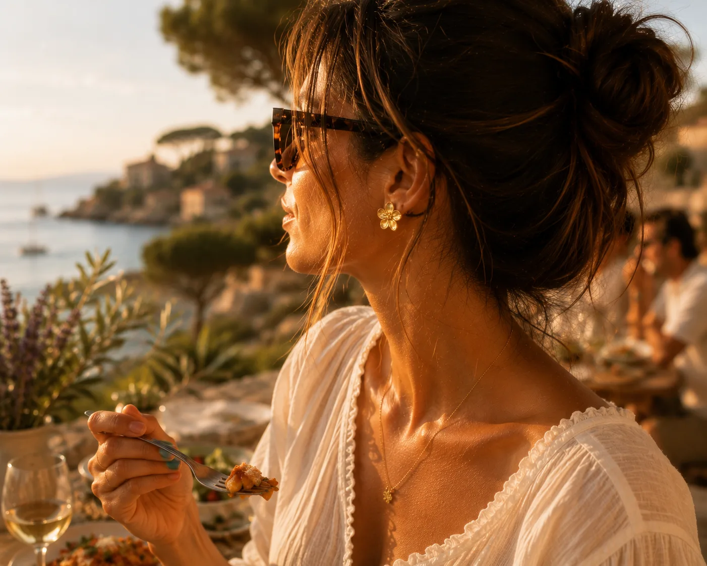 Profil féminin au coucher du soleil, lunettes en écaille, peau dorée, boucle Estrella portée, dîner sur une terrasse en bord de mer méditerranéenne, voiliers à l'ancre