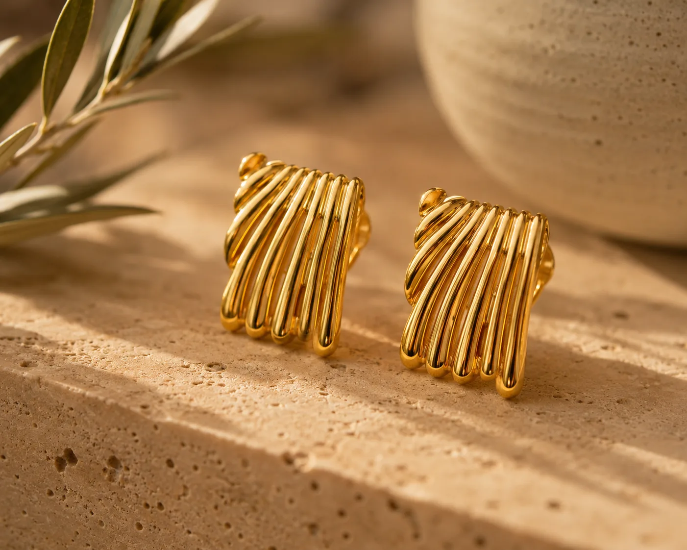 Boucles Duna posées sur un bloc de travertin crème, branche d'olivier à gauche et jarre artisanale en céramique en arrière-plan, lumière dorée