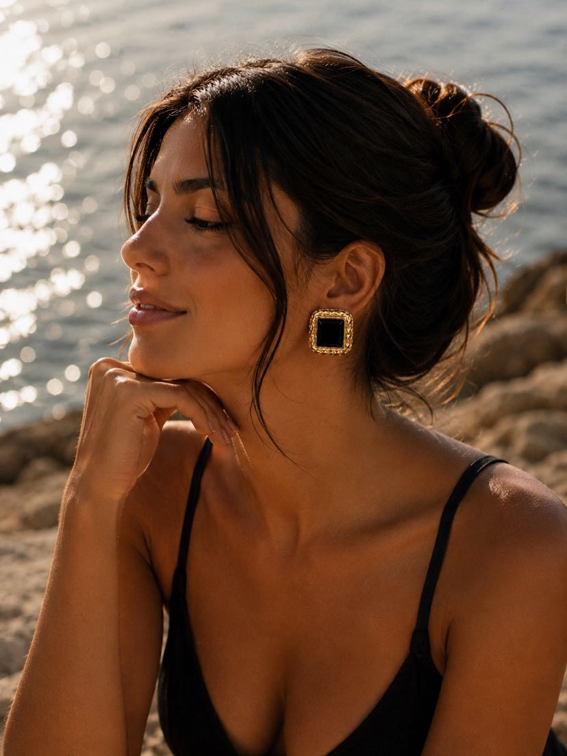 Portrait au bord de la mer, peau dorée, chignon flou, bretelle noire, boucle Paloma carrée or et onyx noir à l'oreille, lumière dorée du couchant sur la mer scintillante