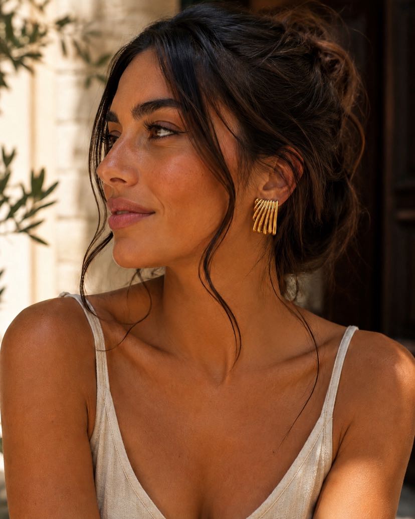 Portrait closeup profil, brune chignon flou et mèches sauvages, boucle Duna golden hour, peau cuivrée, camisole lin écrue, branche d'olivier en arrière-plan